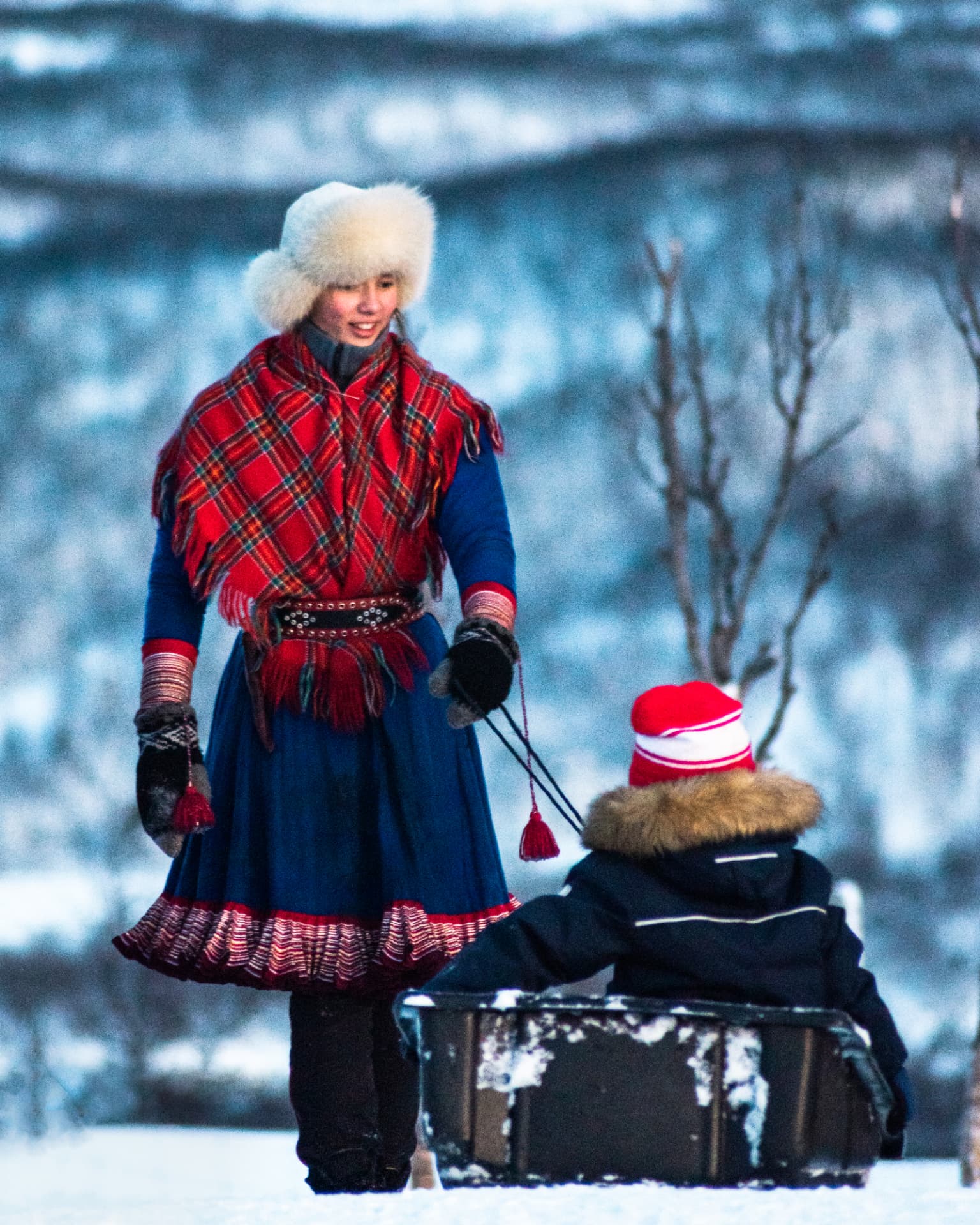 Young family member in gákti pulling a child on a sled through the snow